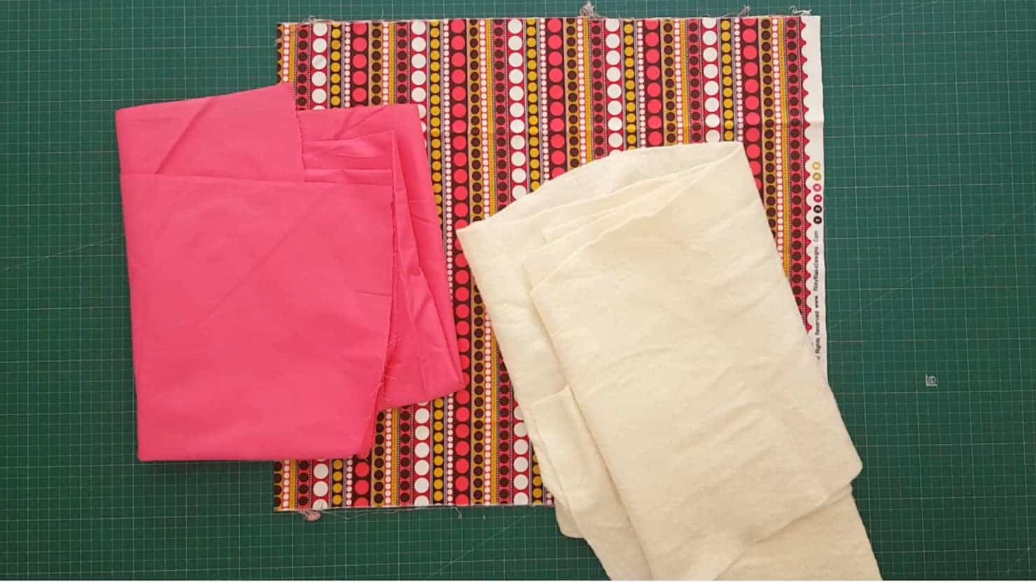 Bright pink and colourful fabric, plus a piece of quilt batting on a cutting mat