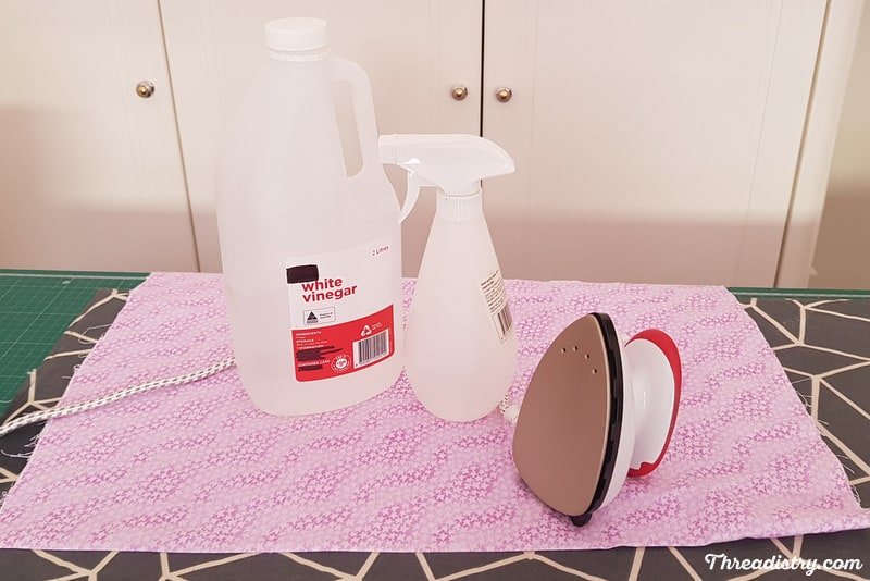 Bottle of white vinegar, spray bottle and iron sitting on creased fabric