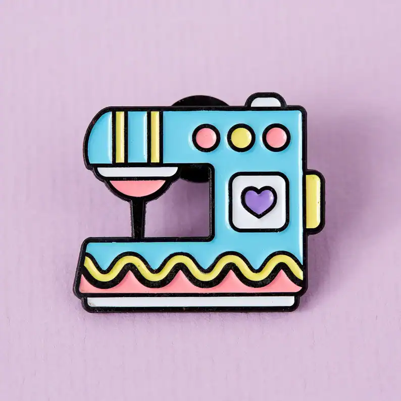 Photography pin badge of a vintage sewing machine with pastel colors, black outline, purple heart button, and pink detail on a pink background.