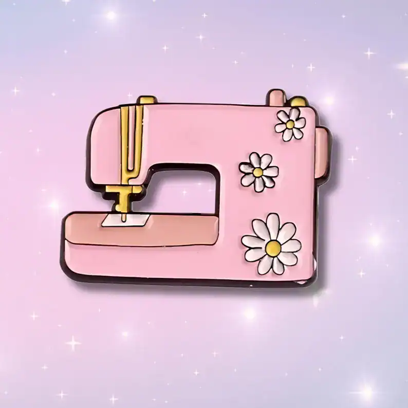 Pink sewing machine shaped pin with daisy flower accents for sewing accessories and crafting enthusiasts.