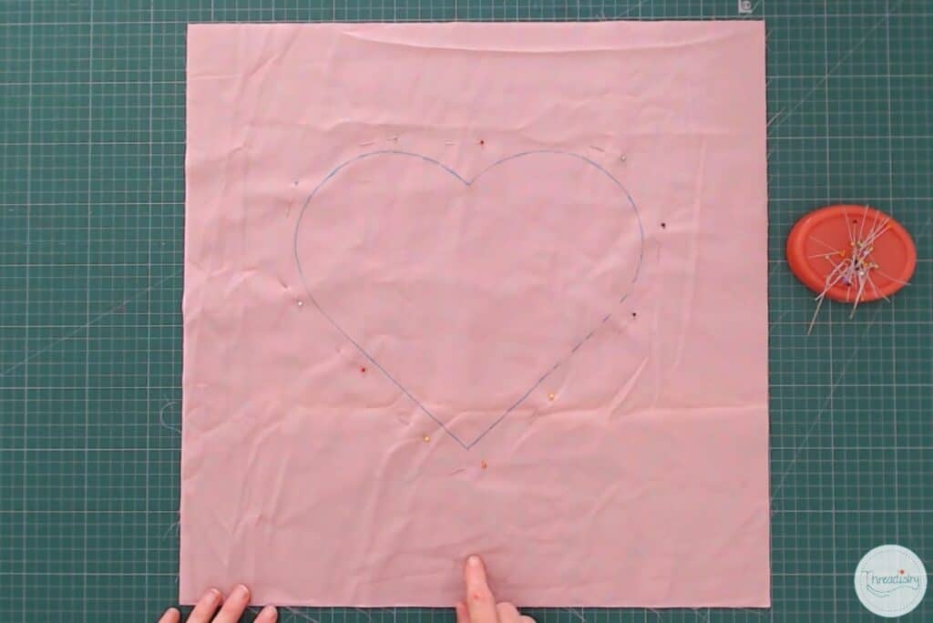Heart drawn with fabric maker on pink fabric, with pins around the heart.