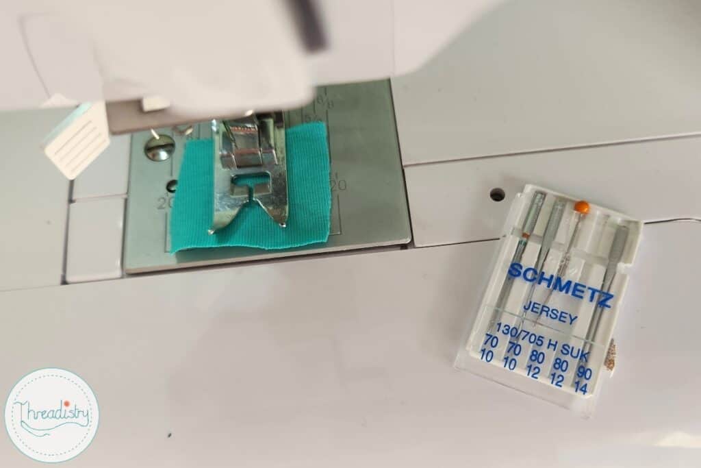 Packet of needles with a pin in one slot sitting on the bed of a sewing machine
