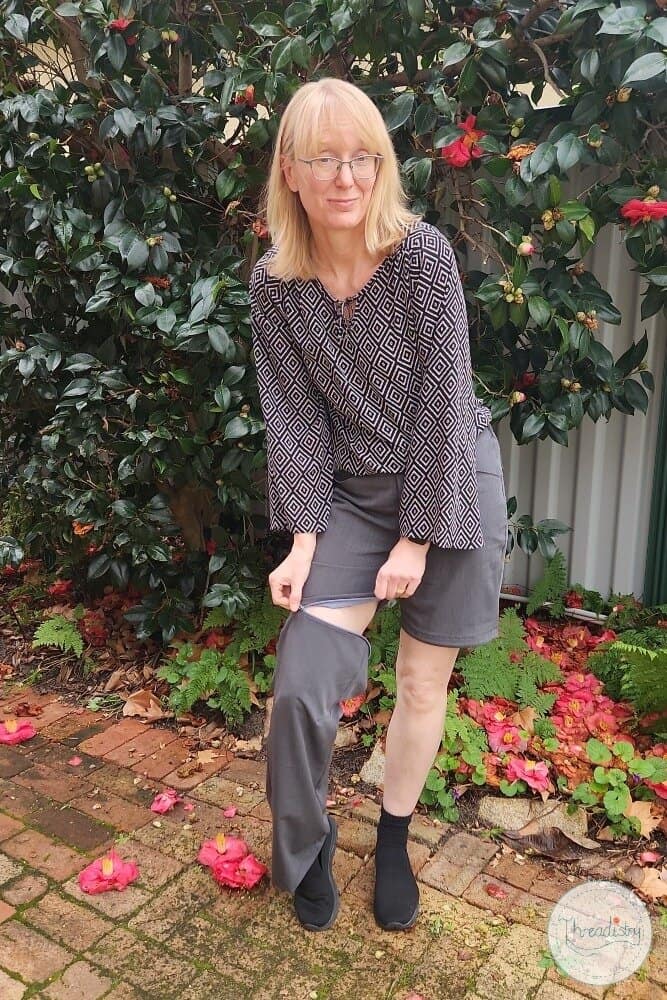 Disoriented woman removing her pants outdoors in a garden with flowering plants and brick pathway.