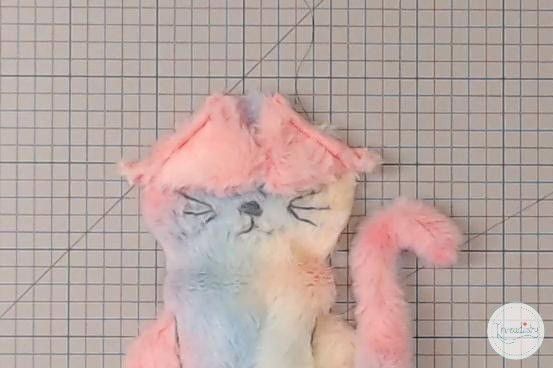 Soft plush cat toy with pink ears and tail, lying on graph paper background.