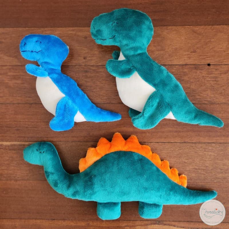 Soft plush T-Rex dinosaur toys in blue, green, and orange with white bellies on wooden surface.