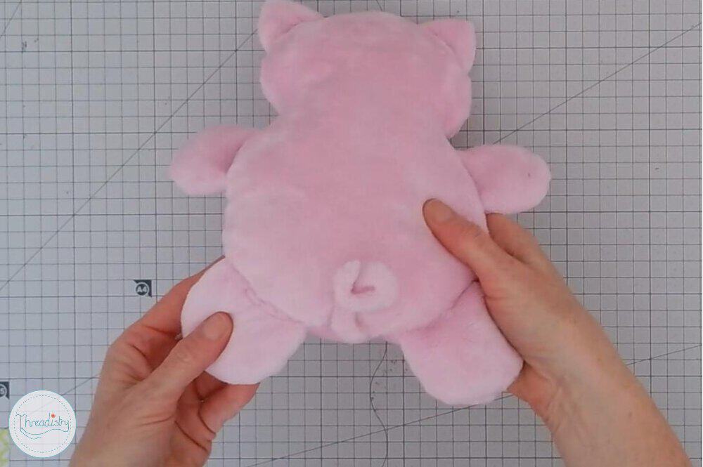 Pink plush piggy toy with a curly tail and small ears on a grid background.