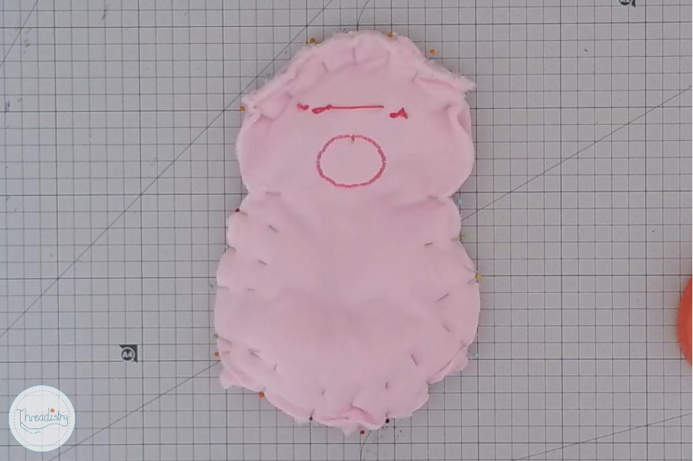 Pink fabric piggy craft with embroidered facial features on grid paper.