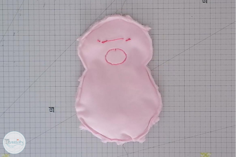 Pink fabric piggy toy with embroidered facial features on graph paper background.