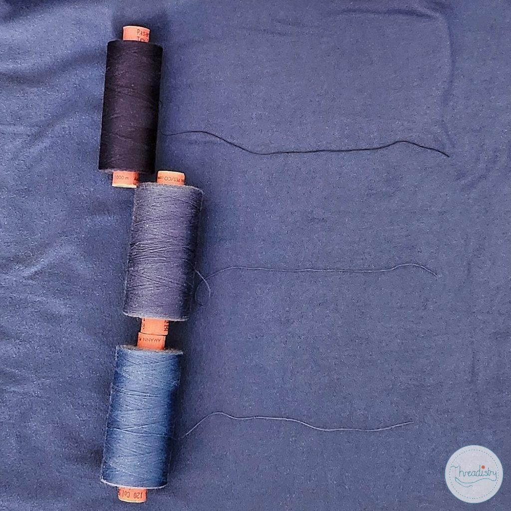 Three spools of thread in black, gray, and blue on dark fabric background.