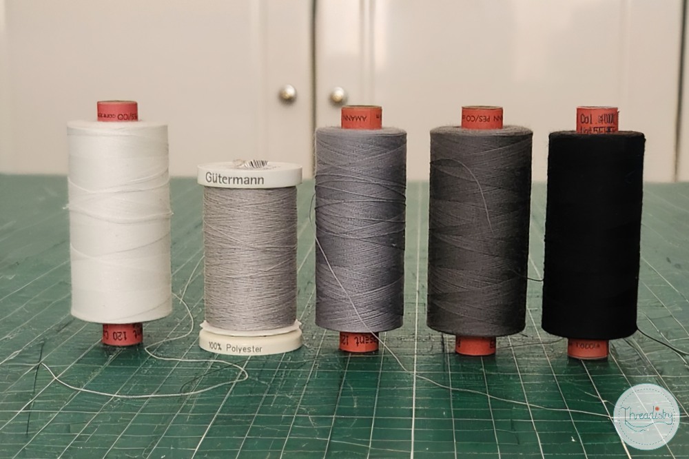 Spools of white, gray, dark gray, and black thread on a green cutting mat.